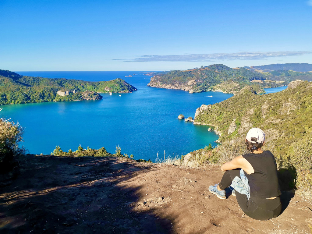 A woman sitting on the top of a hill looking at the blue waters of the Whangaroa Harbour at the top of the Dukes Nose Hike in Northland New Zealand