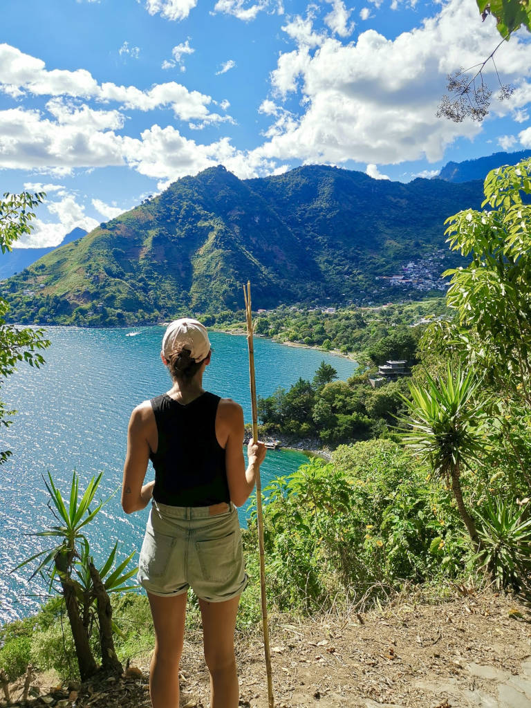 A woman wearing denim shorts a black singlet and a pink hat looking at the view of a beautiful bay while on a hiking trail