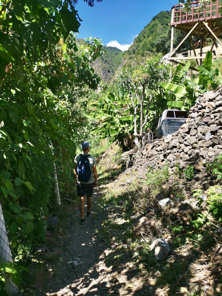 A man with a blue backpack on walking down a narrow dusty hiking trail
