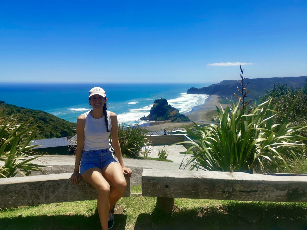 A woman sitting on a wooden bench that has spectacular views of Piha beach in the background