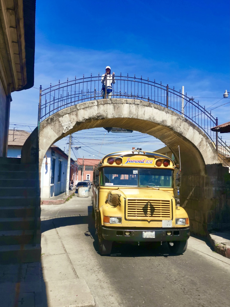 A yellow chicken bus driving under a concrete bridge which is one of the cheapest ways to get from Guatemala City to Quetzaltenango (Xela)