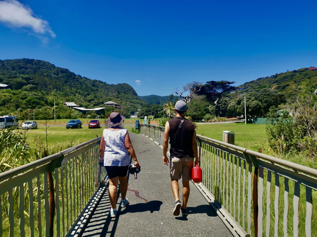 A man and a woman walking along a small bridge at Piha Beach carrying a petrol can on their way to the Mercer Bay Loop Walk