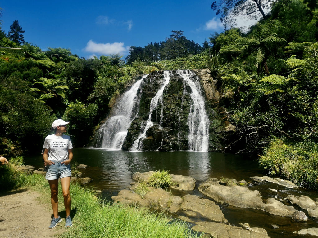 A woman in a white t-shirt and denim shorts standing in front of the Karangahake Gorge waterfall