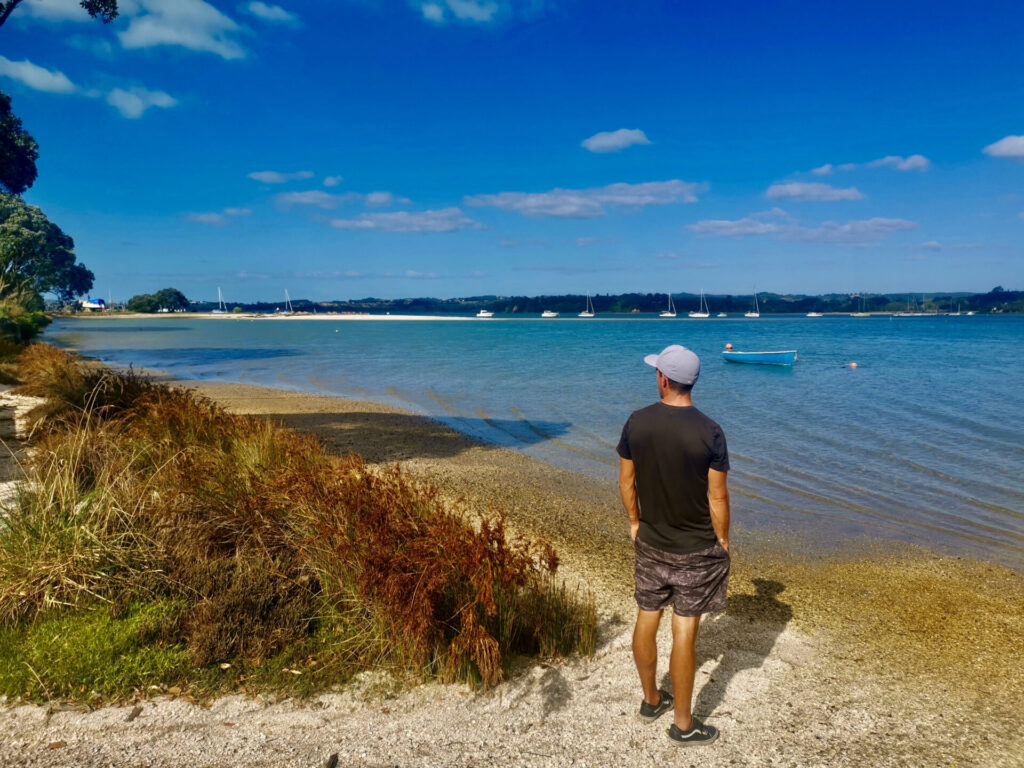 A man in a black t-shirt and shirts standing next to a beach at the end of the Mangemangeroa Reserve walkway one of the best walks in Auckland