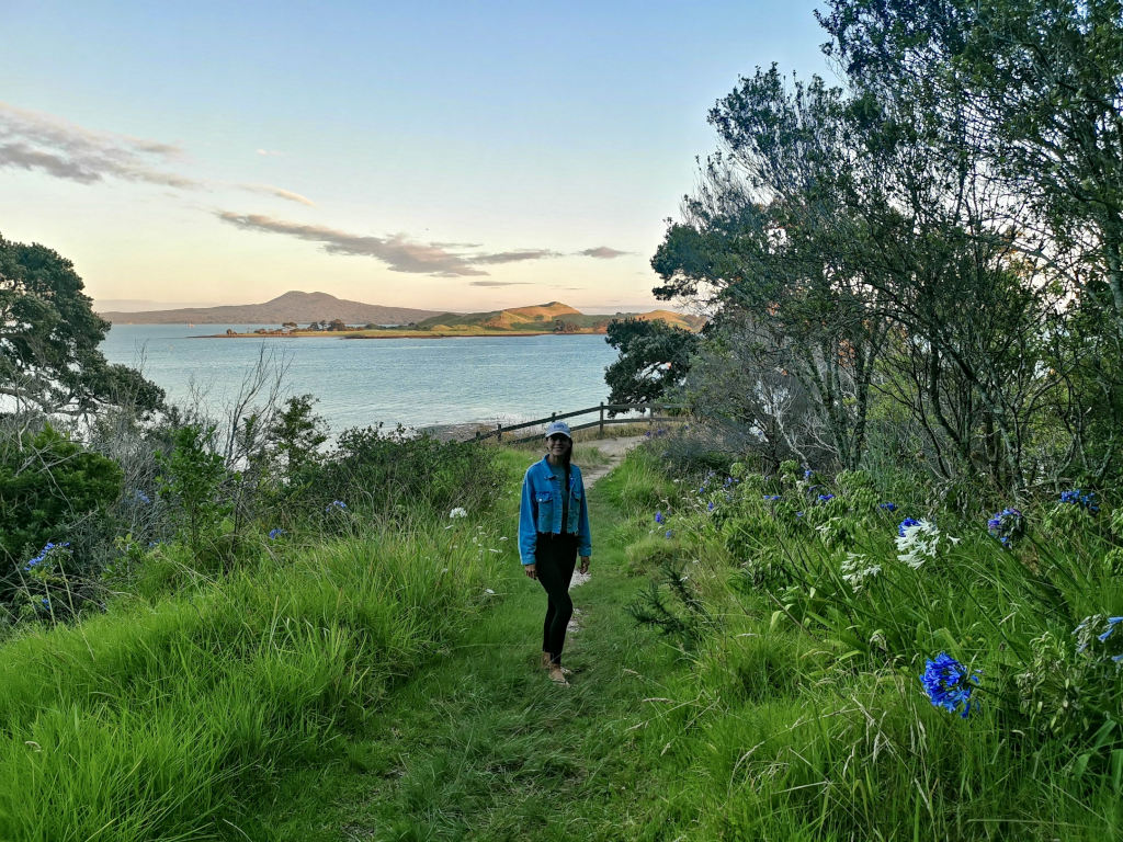 A woman in a denim jacket and black leggings standing on a grassy path with views of Rangitoto island in the background