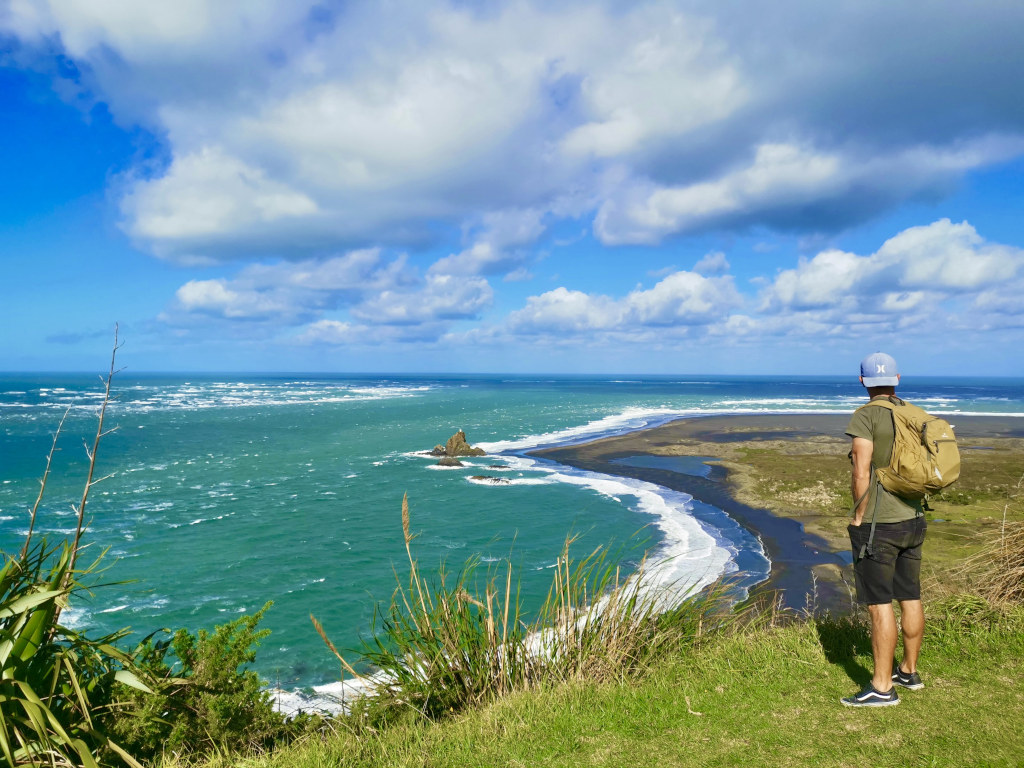 A man with black denim shorts and a backpack on looking out over Whatipu beach from a lookout point on the Omanawanui Track