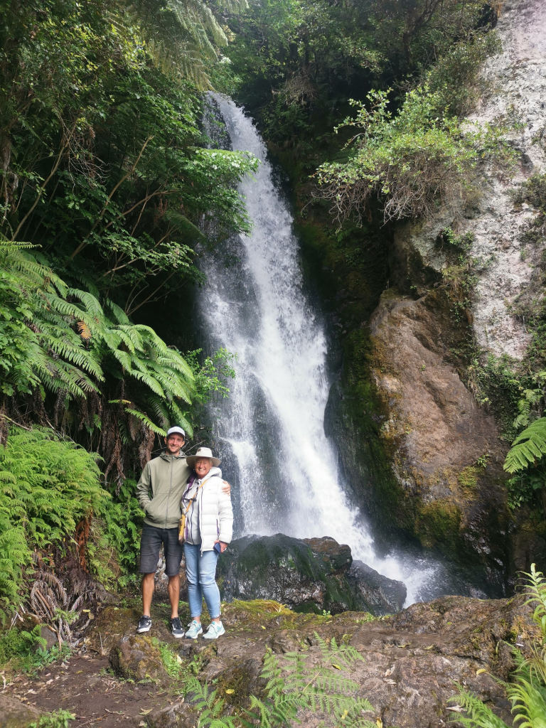 Two people standing with their arms around each other in front a waterfall at the buried village in rotorua