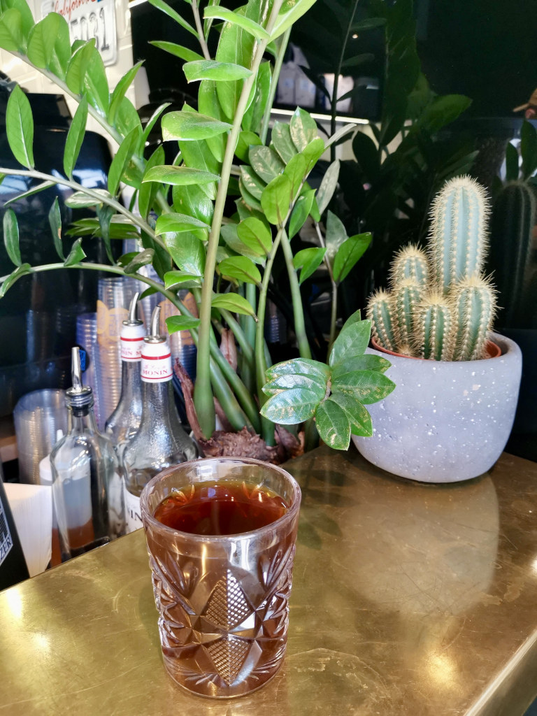 A cup of coffee in a fancy glass sitting on a bench next to a cactus in a small pot plant