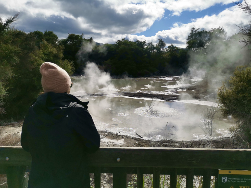 A woman in a pink beanie and a black jacket watching mud pools near the waiotapu thermal reserve in rotorua new zealand