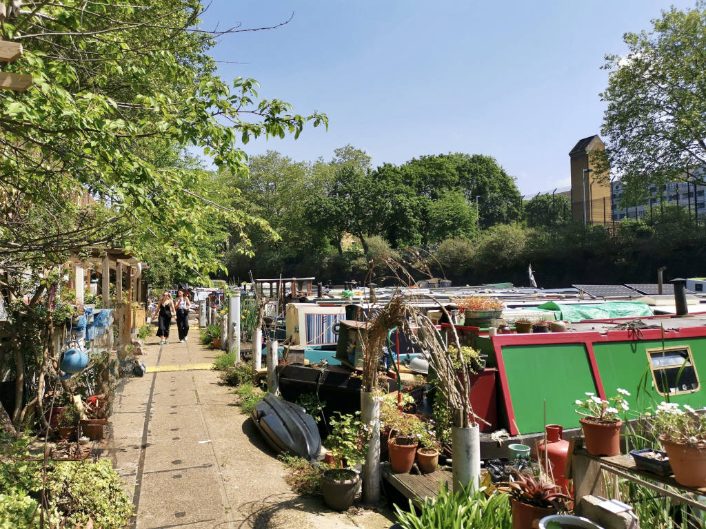 Two people walking along a footpath next to a long row of houseboats on a canal in London