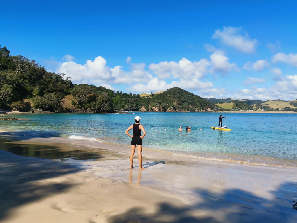 A woman standing at the edge of the wter on a beach with crystal clear water while a man paddle boards just a few meters from shore