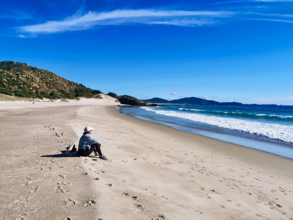 A woman sitting down on ocean beach on the whangarei heads udring a northland road trip