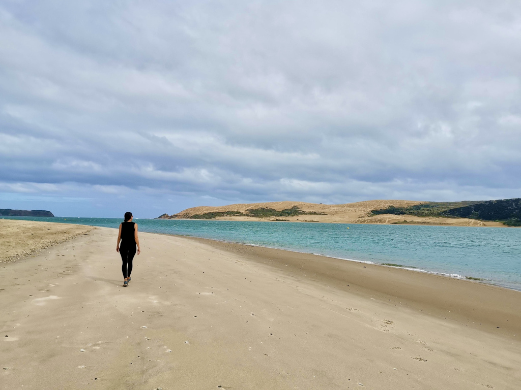 A woman walking down the beach on the edge of a harbour in the distance is a lage sand dune 