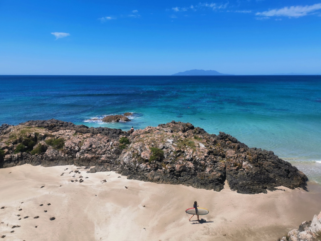 A woman walking down the beach with a surfboard under her arm at tawharanui regional park 