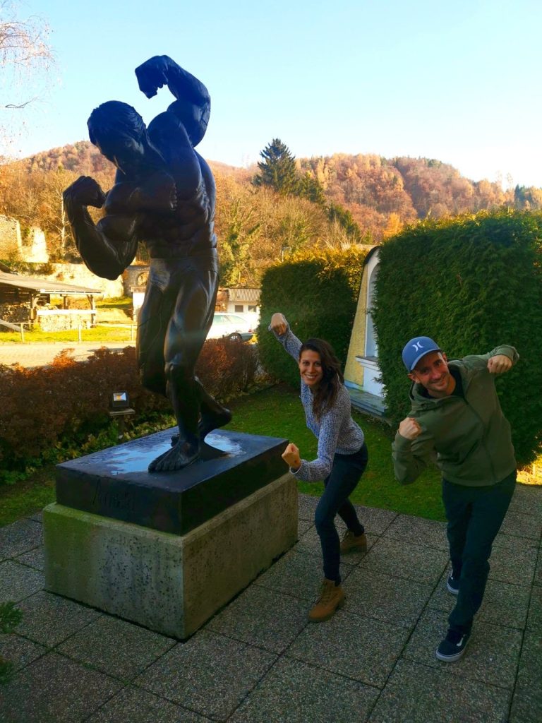 Katharina and Allan posing next to the iconic black Arnold Schwarzenegger statue, mimicking the famous bodybuilder pose by flexing their arm muscles.