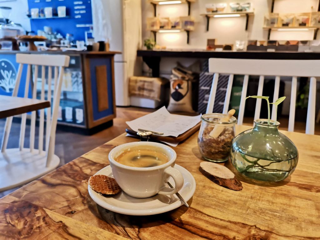 A cup of coffee with dutch stroop waffle on the plate next to the coffee sitting on a table with a vase of water and a clipboard on the table at sango specialty coffee one of the best cafes in amsterdam