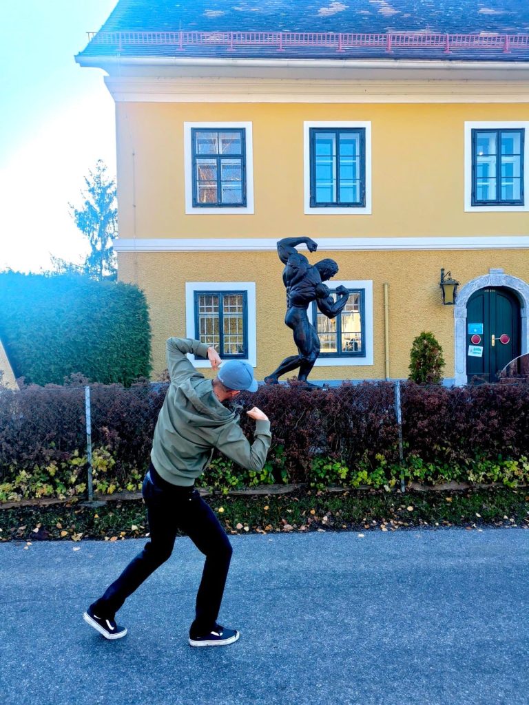 Allan posing in front of the Arnold Schwarzenegger statue in Austria, flexing his arm muscles in a classic bodybuilder pose.