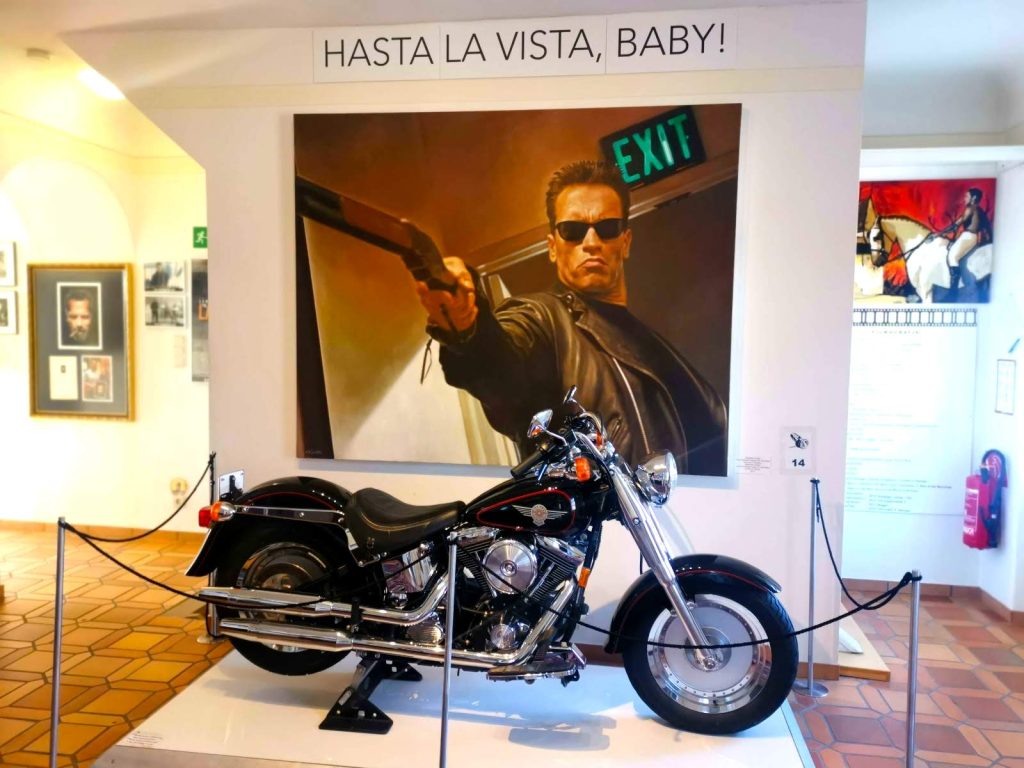 Motorbike from the Terminator movies on a white podium with an Arnold poster saying "Hasta La Vista, Baby" in the back.