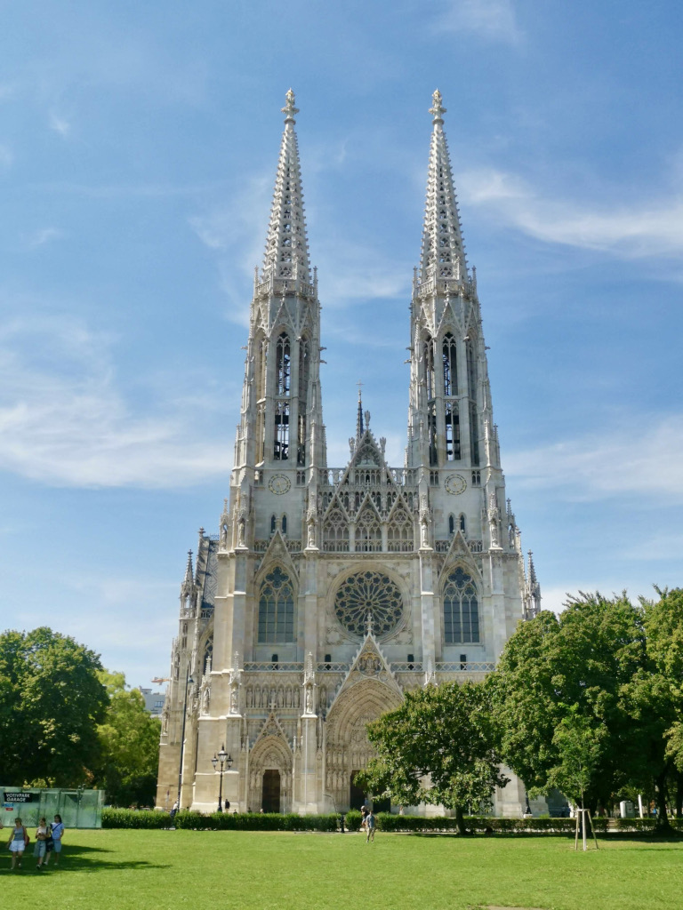 The beautiful votive church a white gothic church with green grass and trees in the park in front of it
