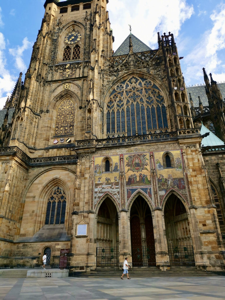 A man in a white t shirt walking past a gothic cathedral at the prague castle during a prague itinerary