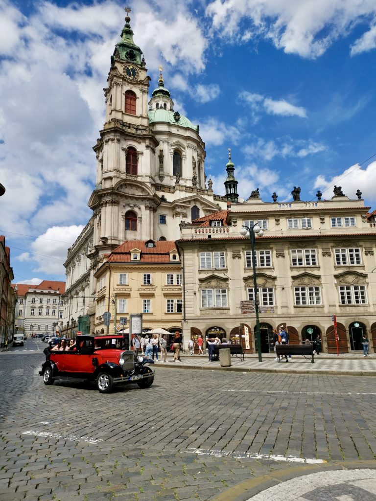 An old red car driving down the road in front of a large church on the streets of Prague