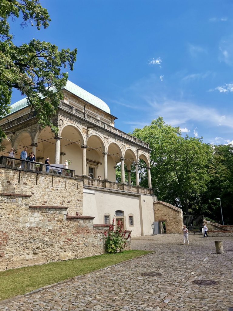 A historical building in a park in prague with tourists walking around in front of it