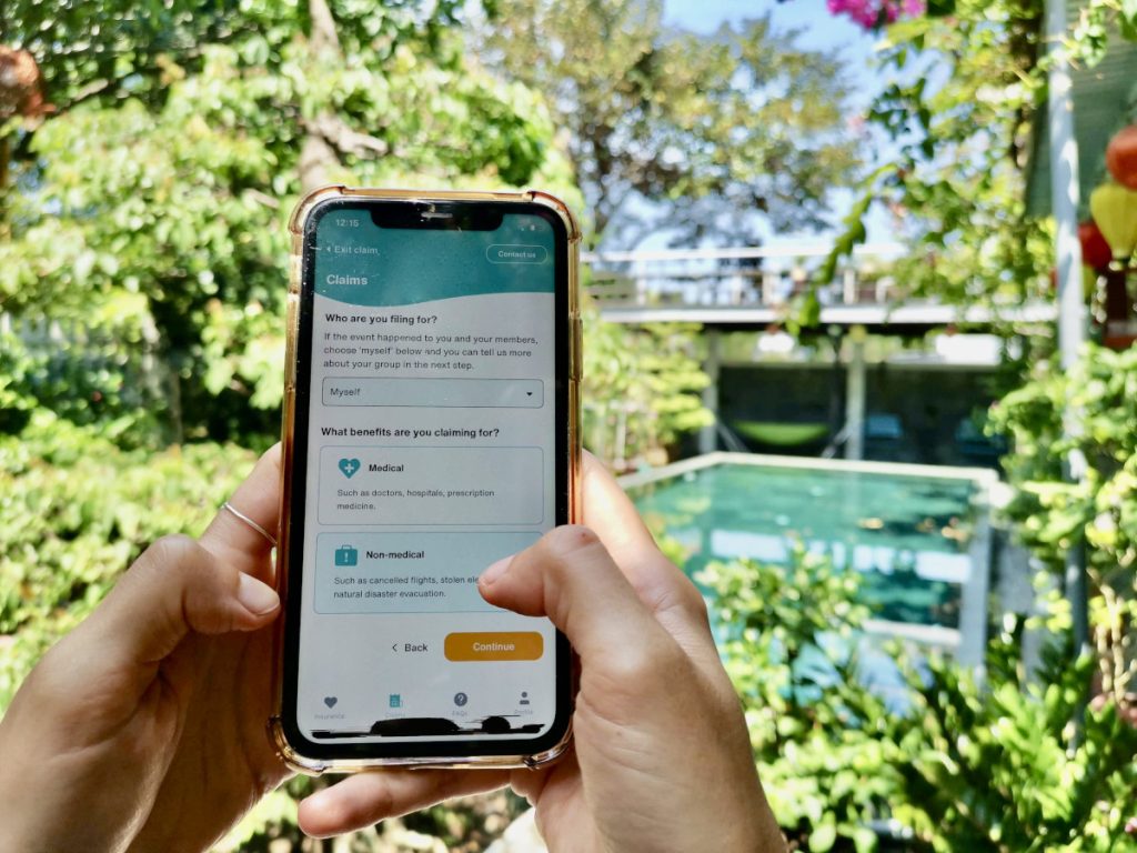 A hand holding a phone next to a pool surrounded by grennery with the SafetyWing App on the screen