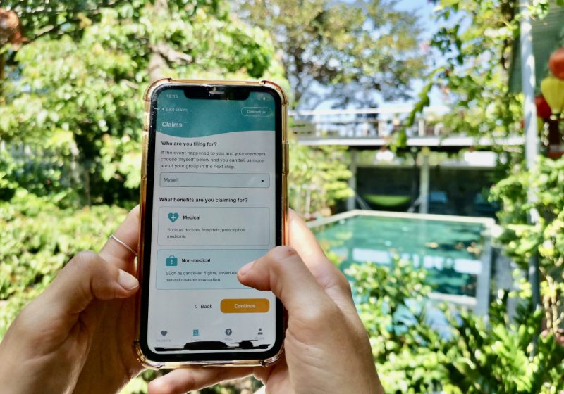 A hand holding up a phone next to a pool surrounded by grennery with the SafetyWing App on the screen.