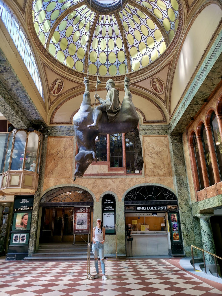 A woman in a white singlet and jeans standing underneath a statue of a man sitting on top of an upside down horse