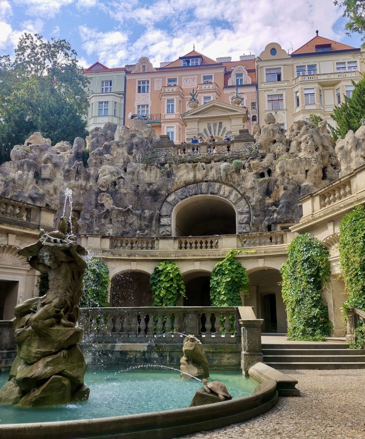 Fountain with a grotto and colorful buildings in the background at Grebovka Park, Vinohrady, Prague.