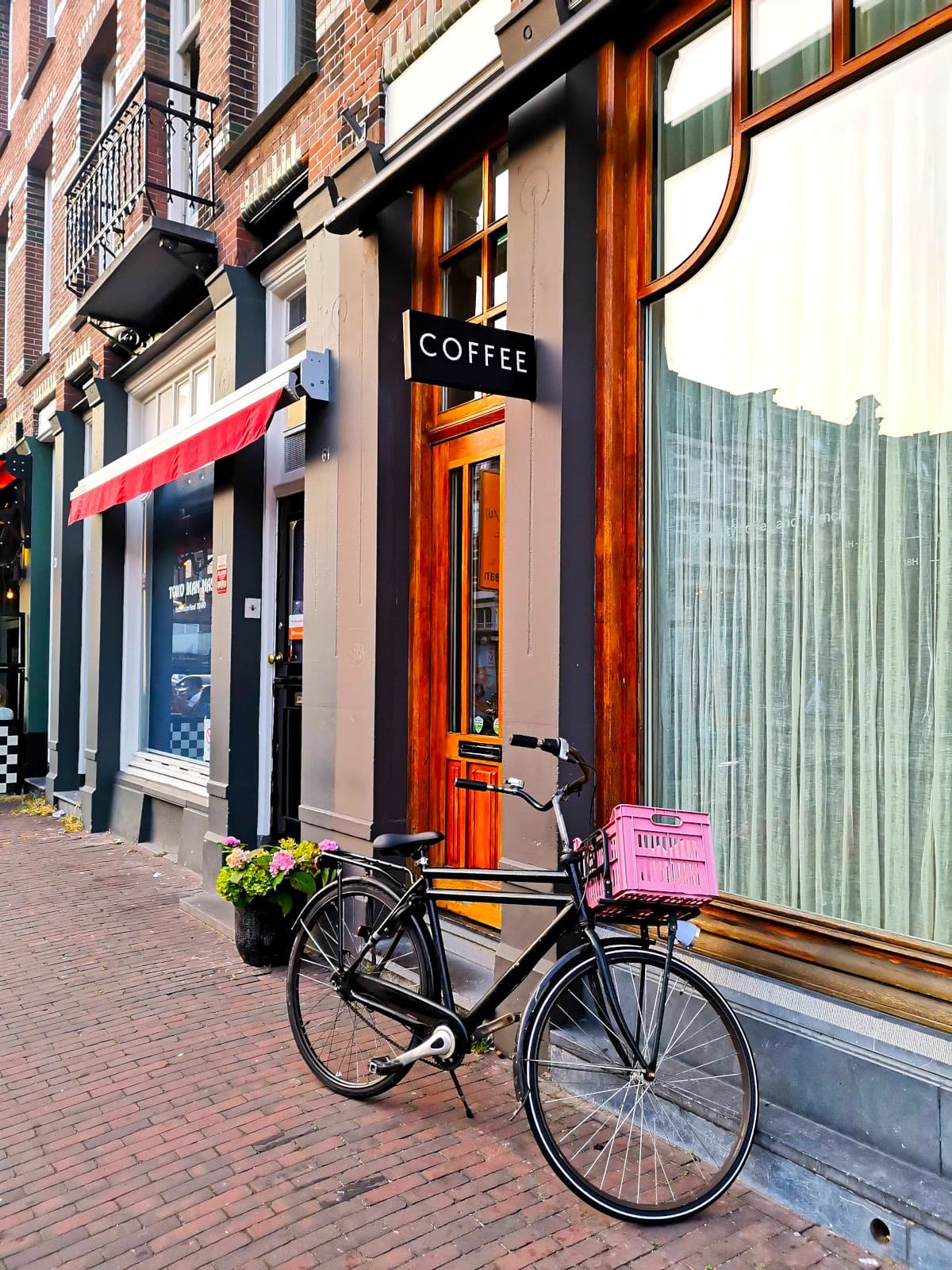 A black bike with a pink basket, parked in front of a typical Amsterdam brick-wall house. The house has a large glass window, a wooden door, and a black sign reading 'Coffee.'