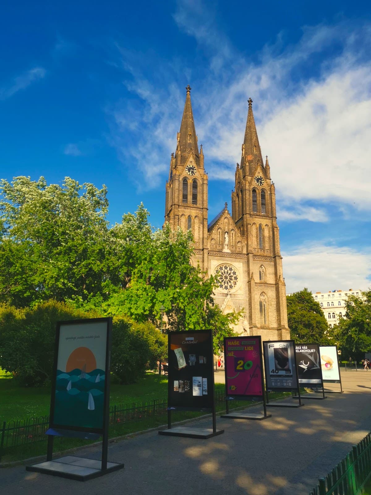 Bazilika sv. Ludmily with two big bell towers surrounded by green trees and signs showing art on a square in Vinohrady.