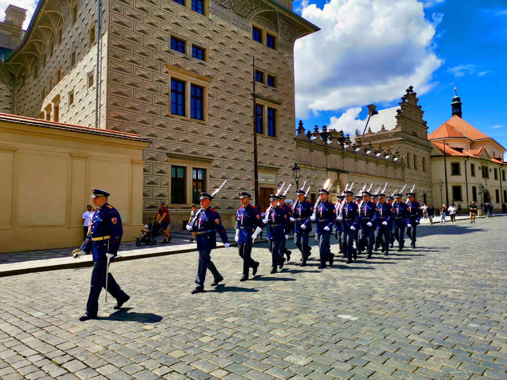 A troop of guards  in dark blue suits marching down a cobblestone street at Prague Castle.