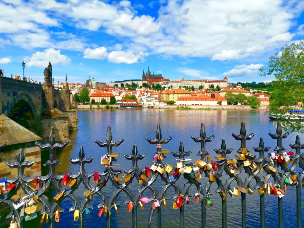 A black metal fence with colorful pad locks attached with the Charles Bridge and Prague Castle in the background.