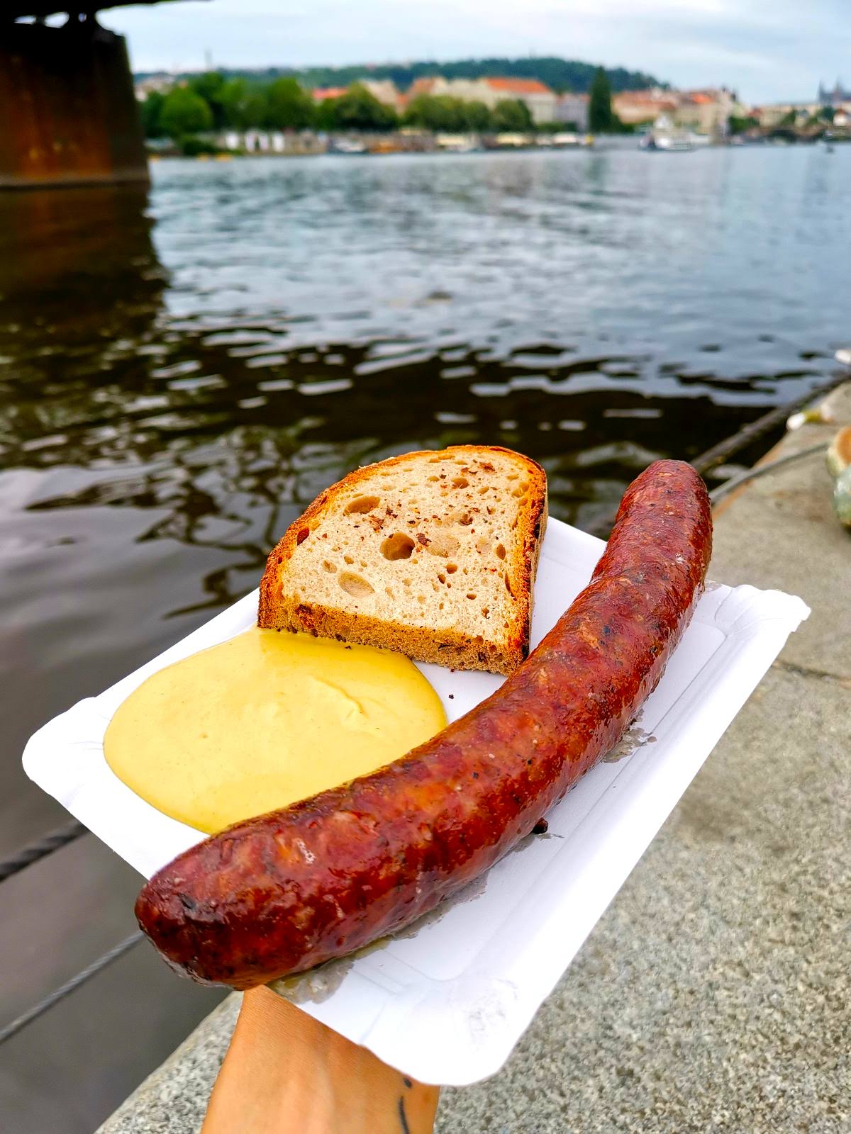 Katharina's hand holding up a white paper plate with a typical Czech grilled sausage, a piece of bread and yellow mustard on it.