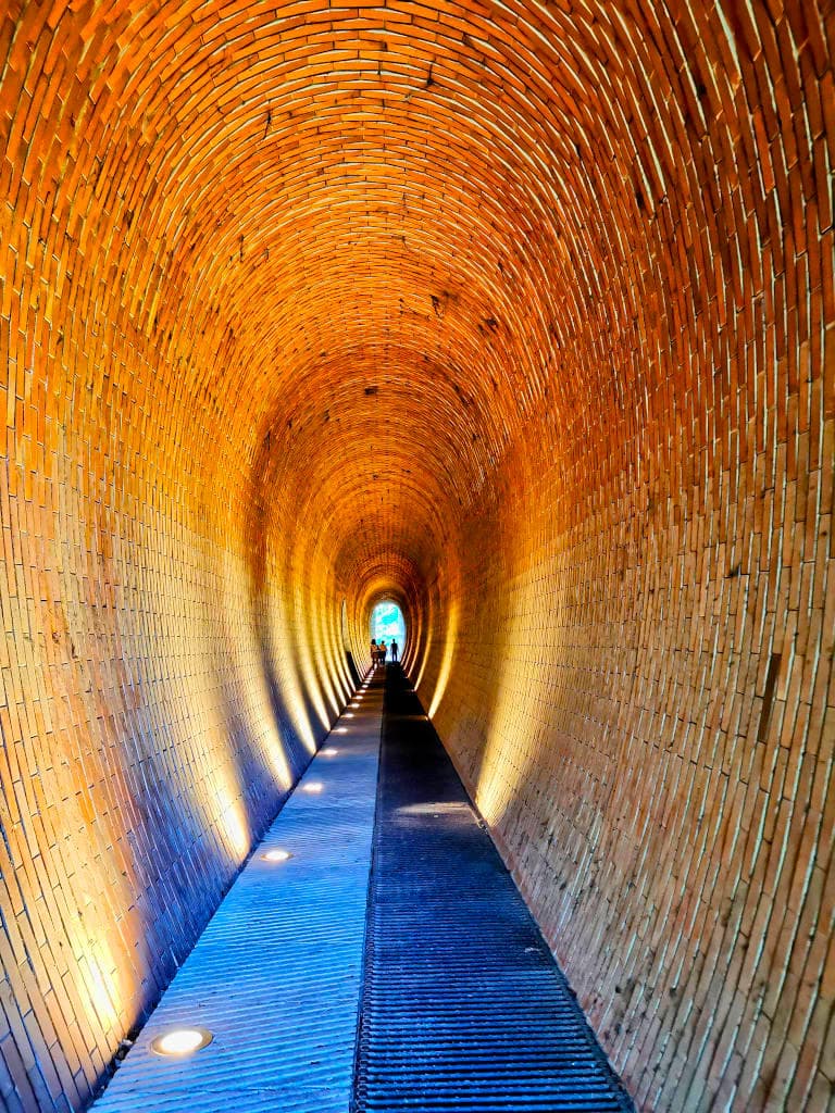 A lit up walk way through a oval shaped tunnel made of bricks in Deer Moat Park, Prague.