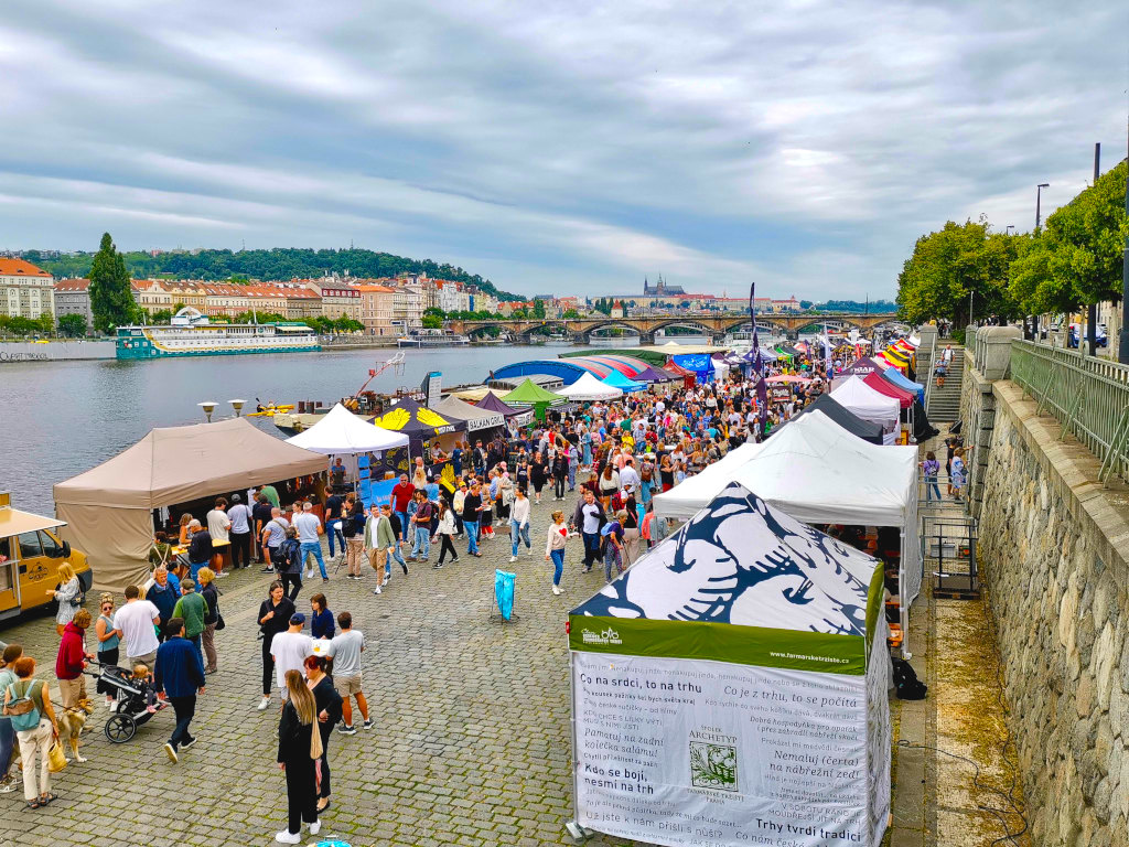 Prague's Farmer's Saturday Market along the Vltava River with colorful tents, a crowd walking and Prague Castle visible in the distance.