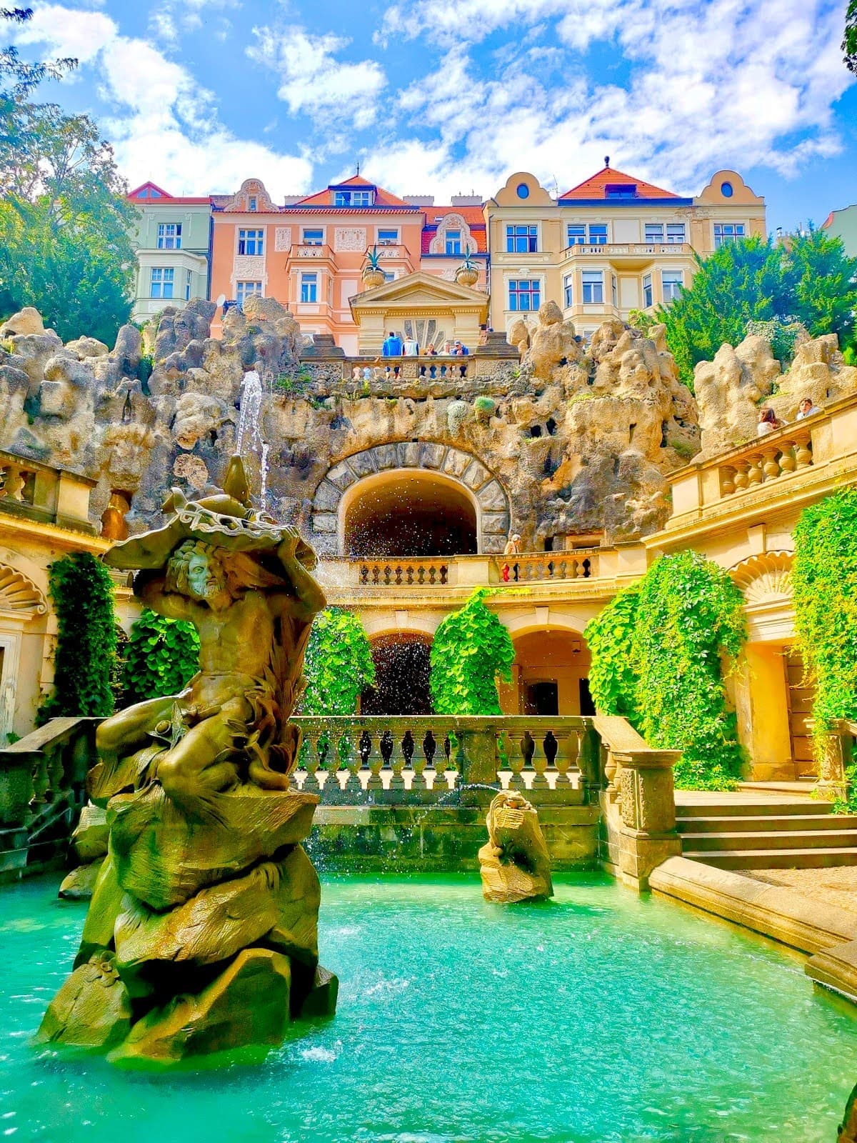 A fountain in front of a man made cave with colorful buildings in the background in Grébovka Park, Vinohrady, Prague.