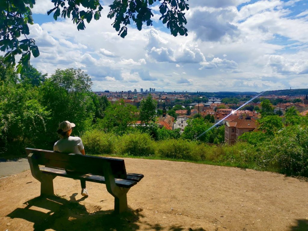 Katharina sitting on a bench in Letná Park overlooking the Vltava River and Prague's red roofs with skyscrapers and hills in the distance.