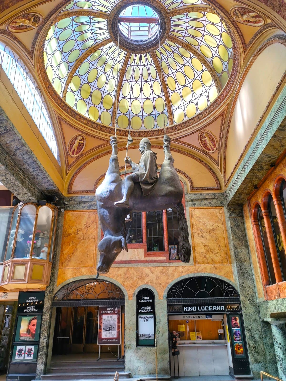 A statue of a man sitting on an upside-down horse hanging from the roof with yellow windows in the Lucerna Passage, one of the hidden gems of Prague.