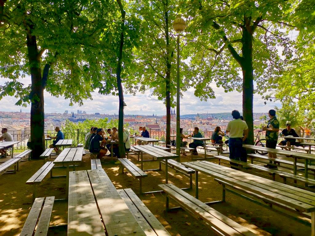 People sitting under trees on wooden benches in a beer garden in Letná Park overlooking the City of Prague.