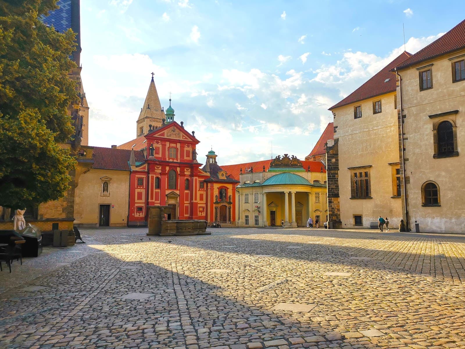 Sunny cobblestone courtyard at Prague Castle with a pink church at the far end.