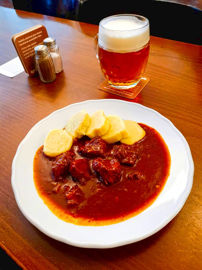 White plate with red traditional Czech goulash with yellow dumplings on a wooden table next to a big jar of beer in a Prague restaurant.