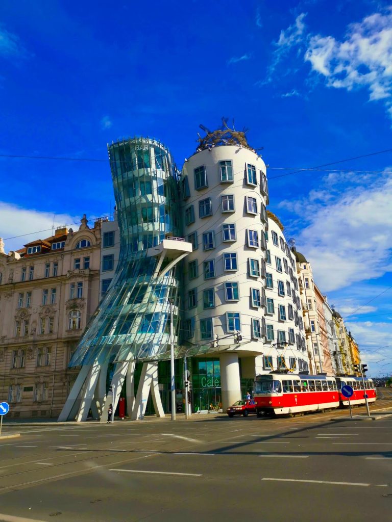 Two white curved houses with many windows known as the Dancing House in Prague with an old-school red tram in front. 