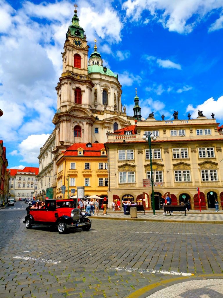 A red oldtimer driving down a cobblestone street in front of a church tower in Prague Malá Strana, a great place to explore on your 2-Day Prague itinerary.