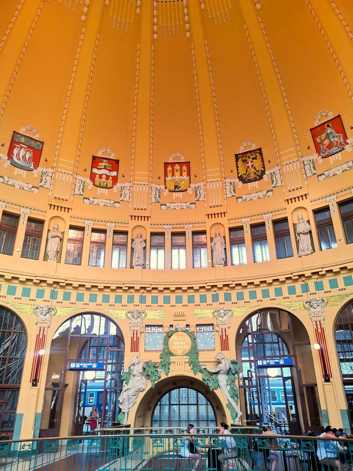 Interior of Prague Main Station's historic yellow Art Nouveau dome with colorful coats of arms, small statues, and architectural details, with people enjoying coffee at Fantova Kavárna below.