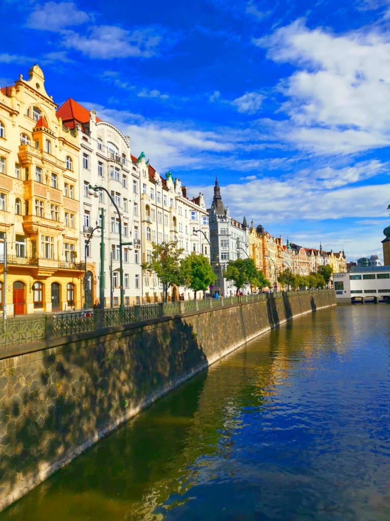 Waterfront of the Vltava River lined with a row of colorful tall Art Nouveau buildings.