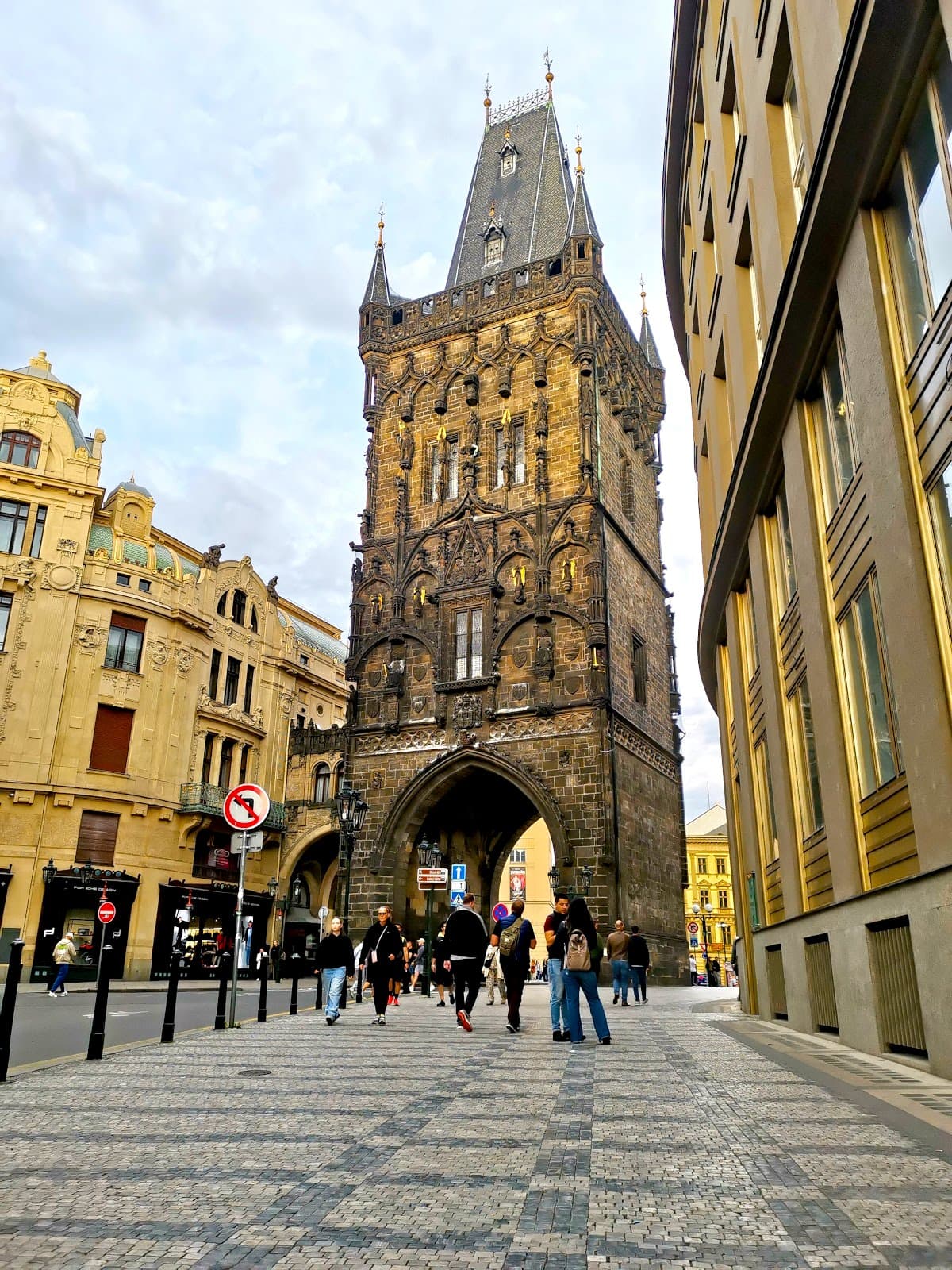 People walking through the Prašná Brána (Powder Tower) in Prague, a historic brown brick tower with a pointed grey roof and a gate beneath.