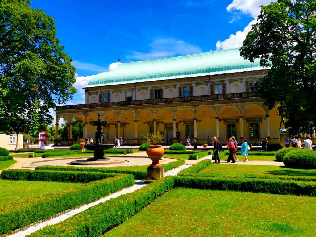 People walking in the sun in front of Queen Anne's Summer Palace with green hedges and a fountain.