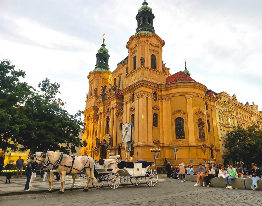 Two white horses pulling a cart in front of the yellow St. Nicholas Church on Prague's Old Town Square.